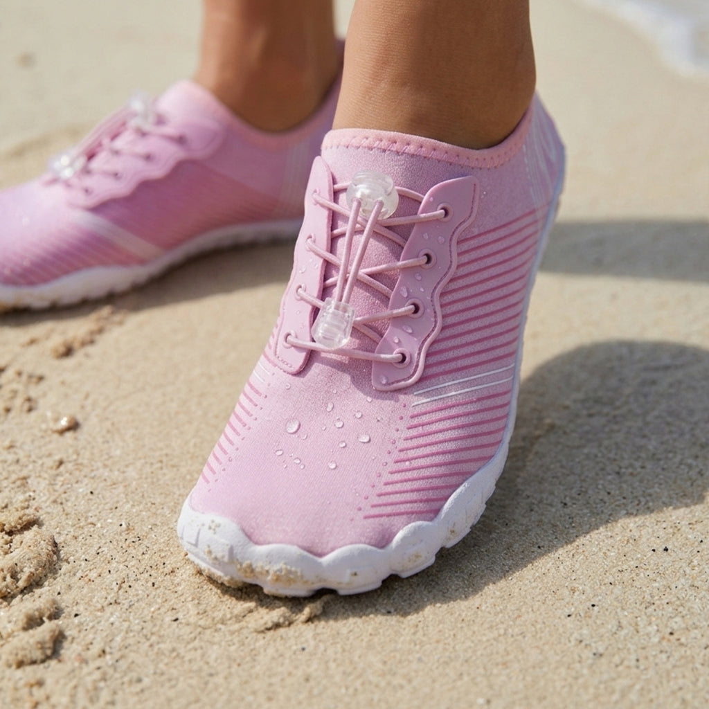 Pink water shoes with clear plastic toggle laces, shown on a sandy beach with water droplets on the uppers.